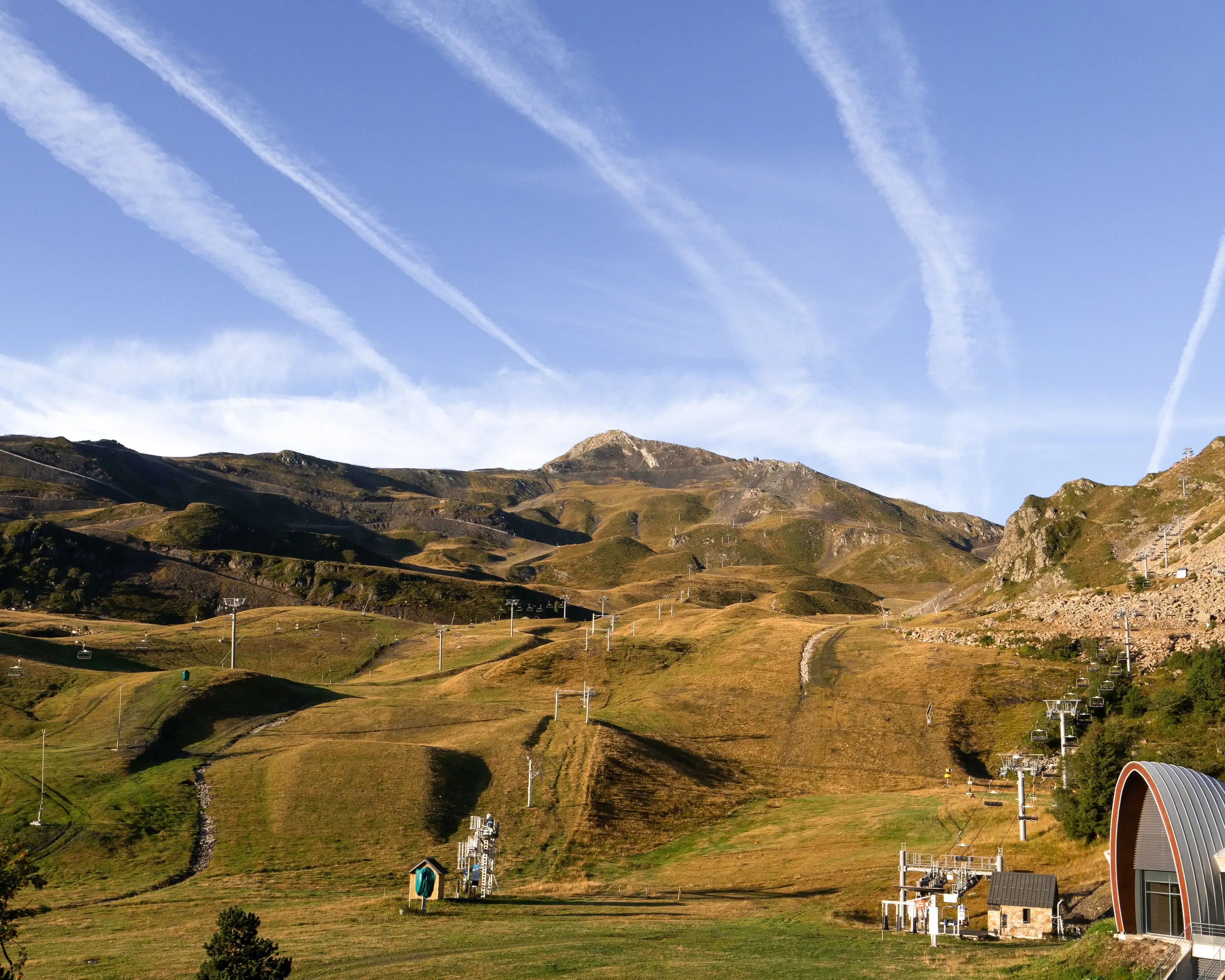 La station de Piau-Engaly aux pentes herbeuses parsemée de remontées mécaniques, face à un sommet sous un ciel bleu traversé par des traînées de condensation.